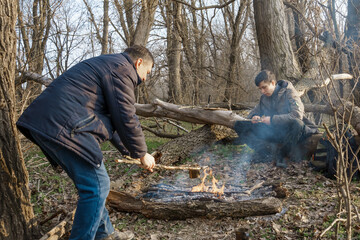 Two men make a campfire for cooking in the forest, father and son sitting on a log while hiking and outdoor activities, early spring landscape