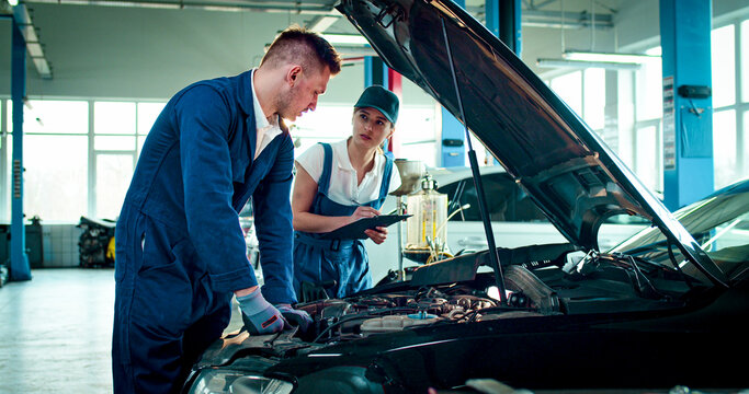 Portrait of two employees wearing specialized clothes work at service station. Young Caucasian people consult each other to fix problems in car.