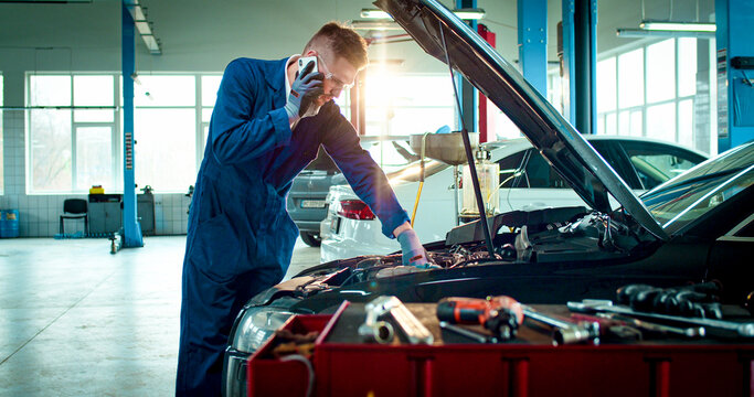 Portrait of young Caucasian male wearing specialized clothes speaks on phone in service station. Handsome man calls customer after repairing car. - Powered by Adobe