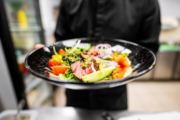 Close-up of a fresh salad with greens, tomatoes, and sliced meat presented in a black bowl held by a person in a black outfit. Perfect for healthy eating, culinary, and lifestyle themes.