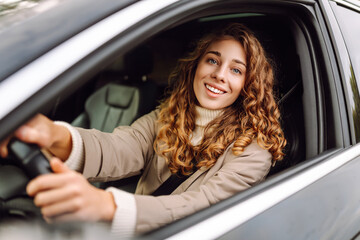 A young woman with curly hair smiling while driving a car in a city during daytime on a clear sunny day