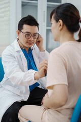 Fototapeta premium Asian female doctor listens to belly of pregnant mother during a prenatal exam in clinic. doctor provides caring advice, ensuring the health and happiness of the expecting mother and baby.