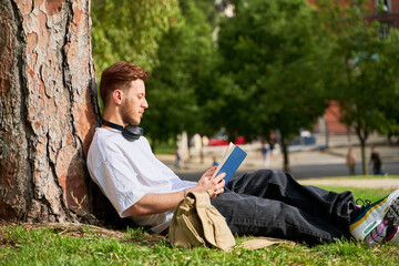 A man is sitting under a tree reading a book. He is wearing headphones and has a backpack next to him. The scene is peaceful and relaxing, with the man enjoying his time outdoors