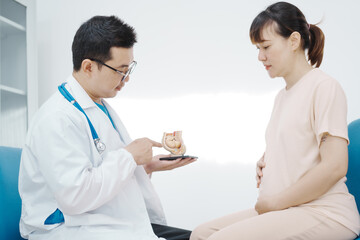 Fototapeta premium Asian female doctor listens to belly of pregnant mother during a prenatal exam in clinic. doctor provides caring advice, ensuring the health and happiness of the expecting mother and baby.