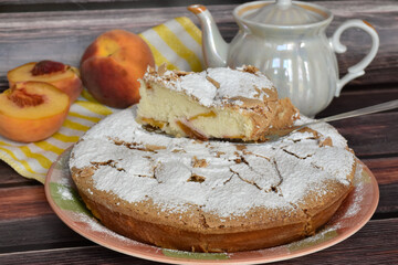Fresh baked homemade charlotte pie sprinkled with powdered sugar. Slice of cake on metal shovel, peach halves, juicy fruit, pearlescent teapot, striped yellow and white napkin on wooden background.