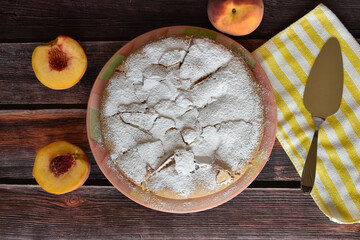 Fresh baked homemade charlotte pie with peaches on wooden table. Cake sprinkled with powdered sugar. Peach halves, juicy peach fruit, metal cake shovel on striped yellow and white napkin.