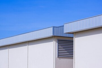 Two industrial warehouse buildings with aluminium roof eaves and rainwater drainage pipelines on white concrete wall against blue clear sky background, perspective side view