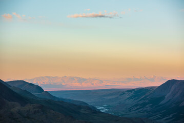 Scenic aerial top view to golden mountain range, illuminated by setting sun under cloud in clear sunset color sky. High mountain ridge far away in soft sunset tones. Gold light on mountain silhouette.