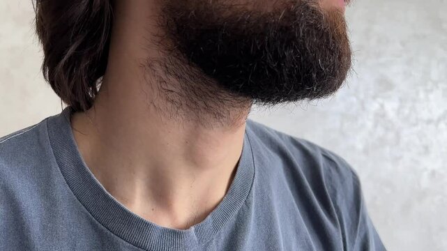 Bearded man in a grey t shirt touching his throat and highlighting his adam's apple in a close up indoor setting