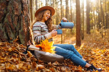 A woman with curly hair enjoys a warm drink while sitting against a tree in a colorful autumn forest during the afternoon