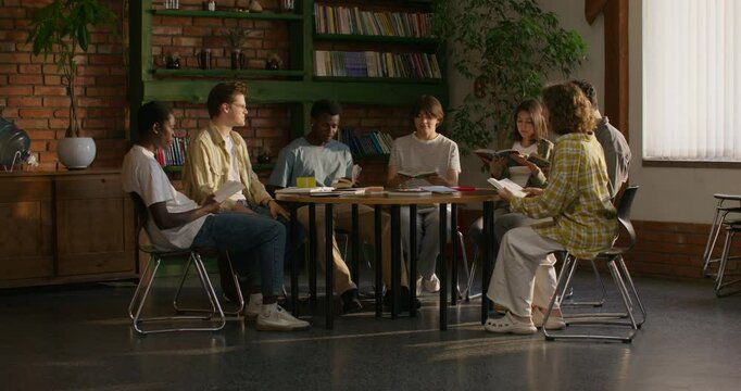 A group of young people of different nationalities join hands and begin prayer while sitting at a table indoors. Daylight