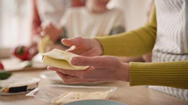 Camera view of woman holding sandwich with one hand. Other hand putting cheese on tasty toast. In background visible family members. People helping mother cooking delicious food. Nutrition concept.