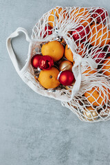 in a white string bag there are tangerines, Christmas tree decorations of red and gold colors, next to it there is a peeled tangerine on a gray background