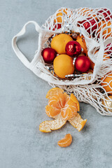 in a white string bag there are tangerines, Christmas tree decorations of red and gold colors, next to it there is a peeled tangerine on a gray background