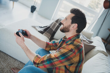 Photo of handsome young guy sit couch playstation video game dressed plaid shirt spend pastime spacious house indoors room