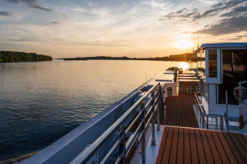 cruiser ship at sunset, chrome railing on sundeck, on river with water, forest, bank in danube...