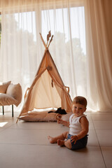 Small child under one year old plays in the room against the backdrop of wigwam in beige shades. The infant baby is happy and dressed  bodysuit. The cozy home environment © Вероника Зеленина
