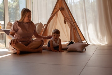 Small child under one year old plays in the room with mom against the backdrop of wigwam in beige shades. The infant baby is happy and dressed  bodysuit. The cozy home environment © Вероника Зеленина
