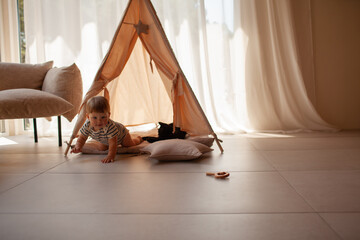 Small child under one year old plays in the room against the backdrop of wigwam in beige shades. The infant baby is happy and dressed  bodysuit. The cozy home environment © Вероника Зеленина