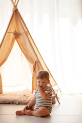 Small child under one year old plays in the room against the backdrop of wigwam in beige shades. The infant baby is happy and dressed  bodysuit. The cozy home environment © Вероника Зеленина