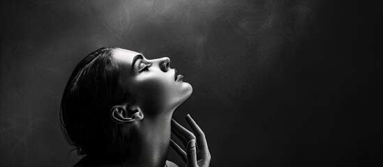 A black and white portrait features a woman tilting her head upwards with her hand touching her neck The dark background produces a dramatic contrast with copyspace above