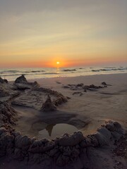 A sand fortress stands proudly on the beach, bathed in the warm glow of the sunset. The soft light highlights the intricate details of the structure, casting long shadows on the wet sand as the waves 