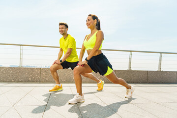 A man and woman performing lunges together on a sunny waterfront promenade, smiling and focused on their exercise.
