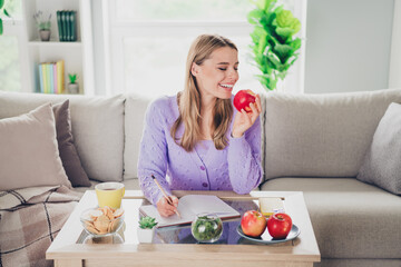 Photo of nice young girl sit couch write notebook table eat bite apple wear violet clothes modern cozy interior flat indoors