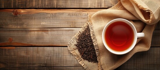 A cup of hot tea rests on sackcloth with dried tea leaves spilling from a bag onto a wooden table providing copy space from a top view It s time to unwind with hot tea in the morning