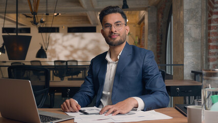 Indian businessman male entrepreneur leader corporate manager work computer laptop write documents paperwork on desk Arabian guy business man happy positive smile guy looking at camera in office cafe