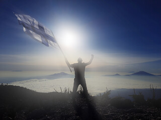 man raising the flag of Finland, on the top of the mountain. background of mountains at sunset or sunrise. Finland flag for Independence Day.