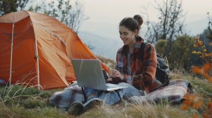 A joyful woman works on her laptop outdoors in a scenic, mountainous area next to a tent, blending work with nature.