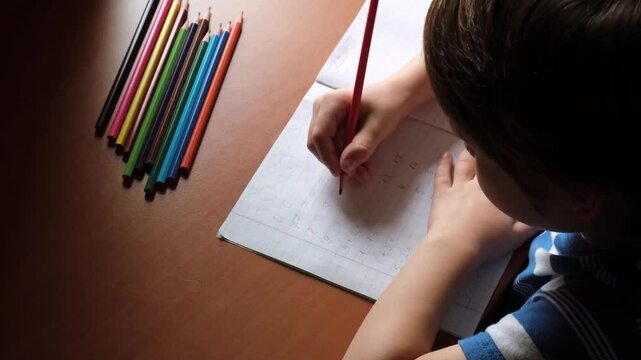 The child writes numbers in a copybook with a colored pencils. A mathematics homework for preschooler. A boy diligently learns to write. The kid is sitting at the table and doing a task. Smart person.