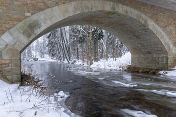 A stone arch bridge with a colourful stream below on a sunny winter day. Lots of snow in the surroundings.
The stone bridge arches over crystal clear water. 
Universal use