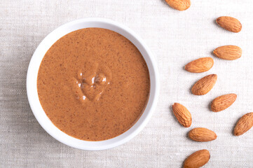 Almond butter in a white bowl on linen fabric. Brown and smooth food paste, made from grinding unshelled almonds into nut butter. Puree of Prunus dulcis nuts with nutty flavor. Good source of protein.