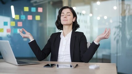 Businesswoman practicing mindfulness meditation at work desk for stress relief. Calm concentrated female office worker in black suit closing eyes and focusing on breath in corporate environment