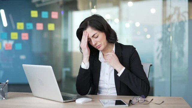 Woman suffering from headache at work. Stress and discomfort evident as she touches her temples, indicating workplace tension or health issues during the workday. Office environment with laptop