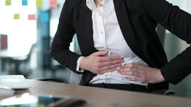 Female hands holding stomach in discomfort, seated at office desk. Businesswoman dealing with abdominal pain during work at workplace in office, suffers from gastritis, poisoning or bloating. Close up
