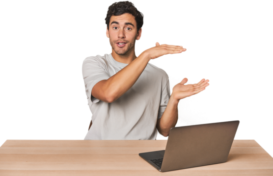 Hispanic young man working on laptop shocked and amazed holding a copy space between hands.