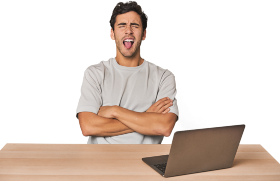 Hispanic young man working on laptop funny and friendly sticking out tongue.