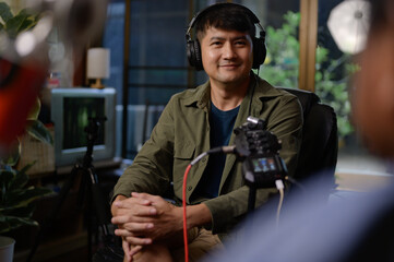 Asian man recording a podcast gestures and smiles as a radio host reads the news into a studio...