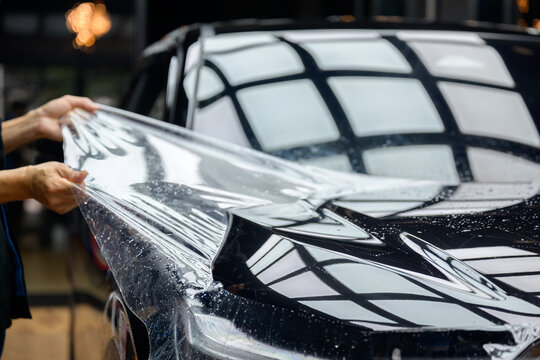 A worker is applying transparent film to a car to protect it from gravel and scratches on the hood of a black car. Applying transparent paint protection film
