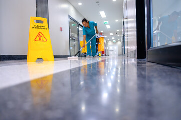 A female cleaner is using a mop to clean the office floor while a cleaning cart and a wet warning...