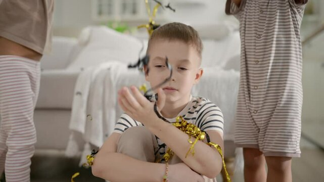 Cute boring five-year-old boy in children party, kids throwing confetti. Bad mood, Caucasian little boy sitting on floor with sad face expression, diverse children playing around, merry Birthday party