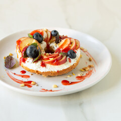bagel with cream cheese, peaches and blueberries on white plate on white table