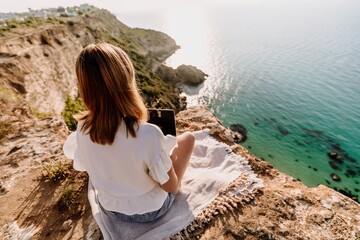 A woman is sitting on a rock overlooking the ocean with a laptop in front of her. She is enjoying...