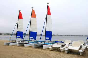 Electric and sail catamaran yachts resting on the beach sand under a gloomy sky over Sail Bay in the early morning, San Diego, California