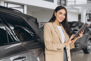 Smiling car seller, businesswoman in beige suit standing in car salon and using tablet for choosing right car.