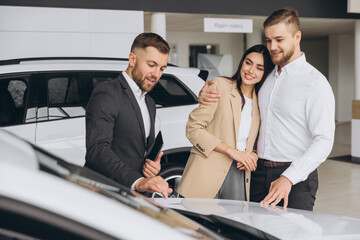 A well-dressed salesman is presenting information to couple in a car dealership showroom. The couple appears interested as they stand between two new vehicles