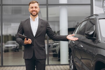 Fototapeta premium Portrait, smiling man at outside car dealership for car sale in a commercial parking. Business, luxury and automobile trade with a happy bearded salesman in suit outdoor for transport service
